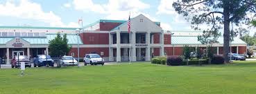 Picture of The Atmore Community Hospital. There is a parking lot out front with cars, a flag pole, The United States flag, an open field, bushes, and a tall pine tree